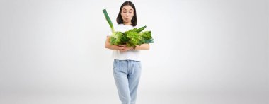 Sağlıklı vejetaryen yaşam tarzı. Mutlu Asyalı kadın en sevdiği sebzelere bakar, elinde lahana, marul ve yeşil organik gıda, beyaz arka plan.