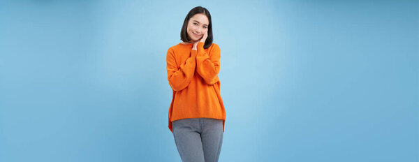 Красота и благополучие. Молодая брюнетка смеется и улыбается, трогает свое естественное здоровое лицо, стоя на синем фоне студии.