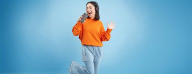 Güzel Koreli kız akıllı telefon mikrofonuyla şarkı söylüyor, kulaklıkla müzik dinliyor, mobil uygulamayla karaoke yapıyor, mavi arka planda duruyor..