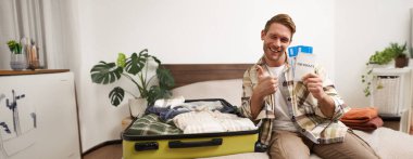 Mutlu, heyecanlı genç adamın portresi, pasaportunu ve uçak biletlerini tutuyor, giysilerini bavula koyuyor, yaz tatiline hazırlanıyor, tatile çıkıyor ve heyecanlanıyor..