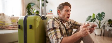 Şok olmuş suratlı bir adam, bavuluyla yerde oturuyor, endişeli ve cep telefonu ekranında gergin görünüyor. Boşluğu kopyala