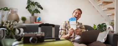 Mutlu yakışıklı adamın portresi, uçuş biletlerini gösteriyor, yaz tatili için yer ayırtıyor, dizüstü bilgisayarla oturma odasında oturuyor, kameraya gülümsüyor..