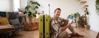 Yakışıklı karizmatik bir adamın portresi, bavullu bir turist, yere oturuyor, pasaport ve bilet tutuyor, göz kırpıyor ve kameraya gülümsüyor..