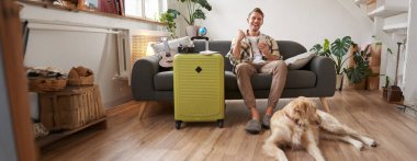 Heyecanlı bir adam, köpekli bir turist, valiz ve seyahat kamerasıyla oturma odasında oturuyor, adam evet diyor ve kutluyor, internetten fiyatlardan memnun, hayvan dostu otel rezervasyonları yaptırıyor..