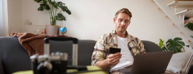 İnternetten bilet alan neşeli bir adamın portresi dizüstü bilgisayar ve kredi kartıyla otel rezervasyonu yapan oturma odasında bavulun yanında oturan....