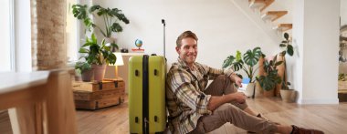 Gülümseyen yakışıklı adamın portresi, bavuluyla yerde oturuyor, elinde pasaport ve uçak biletleri var, turneye çıkıyor..