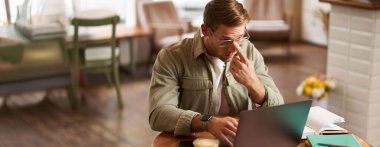 Genç dijital göçebe imajı, gözlüklü adam kafede oturuyor, kahvecide çalışıyor, iş yerinde dizüstü bilgisayar kullanıyor, gözlük takıyor, içkisini içiyor..