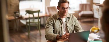 Uzaktan çalışma ve serbest çalışma kavramı. Gözlüklü yakışıklı adam dizüstü bilgisayar ve dizüstü bilgisayarla oturur, online çalışır, kahve dükkanından uzaktan çalışır, bilgisayarla kafede oturur..