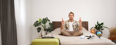 Yaz tatili için bavulunu topladıktan sonra yatakta meditasyon yapan genç bir adamın portresi, bacak bacak bacak üstüne atmış, nirvana pozu veren, rahatlatıcı, bir şeyler umut eden. Turizm ve farkındalık kavramı