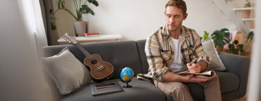 Kontrol listesi yazan, tatil için eşyalarını toplayan, notlarıyla karşılaştıran, dünya ve ukulele ile oturma odasında oturan adam..