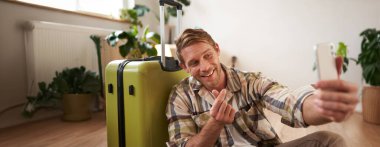 Gülümseyen yakışıklı adamın portresi, selfie 'de parmak izi, cep telefonunda kendi fotoğrafını çekiyor, bavulla poz veriyor, tatile çıkıyor. Turizm konsepti