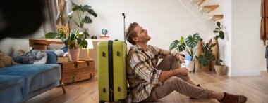 Erkek modelin bavullu portresi, yerde oturuyor, uçak biletleri ve pasaport tutuyor, yurtdışına gidiyor, havaalanına gitmek için taksi bekliyor..