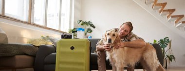 Köpeğini kucaklayan, evcil hayvan dostu bir otele tatile giden, bavulunu toplayan, oturma odasında oturan genç bir adamın portresi..