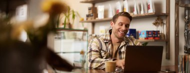 Kafede dizüstü bilgisayar kullanan yakışıklı bir gencin portresi, yaratıcı kamusal alanda bilgisayarla oturuyor, mutlu bir gülümsemeyle klavyeye yazı yazıyor..