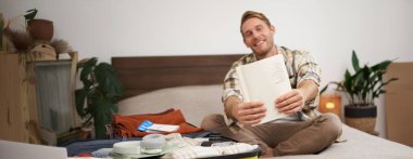 Gülümseyen, yakışıklı bir adam portresi, bavula bir şeyler koyması, defterde yazılı listesini göstermesi, tatile çıkması. Turizm ve yaşam tarzı kavramı