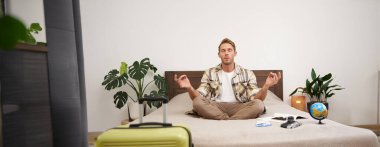 Düşünceli genç bir yolcunun portresi, yatakta oturup meditasyon yapan, bavulunu toplayan, yurtdışına seyahat eden, yoga yapan, yanında bir küresi ve kamerası olan bir adam..