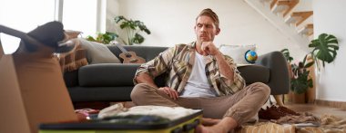 Düşünen ve bagajına bakan bir adamın portresi, açık bavuluyla oturuyor, tatil için hazırlanıyor. Turizm, seyahat ve insanlar konsepti