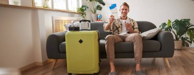 Mutlu adamın portresi internetten akıllı telefondan alınmış, elinde biletler, bavulun yanında oturuyor, tatile çıkıyor, tatile hazırlanıyor..