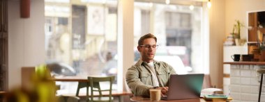 Ciddi yüzlü bir adamın portresi, kafede oturuyor, gözlük takıyor, dizüstü bilgisayarda çalışıyor, konsantre görünüyor, serbest çalışıyor..