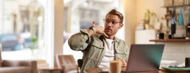 Gözlüklü genç yakışıklı bir adamın portresi, kafede çalışan iş adamı, dizüstü bilgisayarla oturan, telefonla el hareketi gösteren, servisini tanıtan.