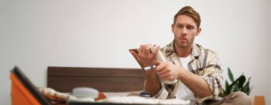 Elbiseleriyle valizine ciddi şekilde bakan genç bir adamın portresi, kontrol listesini gözden geçiriyor, elinde notlarla defteri tutuyor, tatilde ne getireceğini seçiyor. Turizm konsepti.