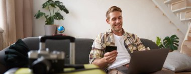 Kredi kartı ve dizüstü bilgisayarıyla gülümseyen yakışıklı adam, tatil için otel rezervasyonu yapan turist, valizin yanında oturuyor..