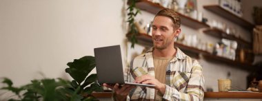 İnternette çalışan genç bir adamın portresi, kafede oturan, dizüstü bilgisayarda projesini yapan, kahve içen, ekrana bakarken gülümseyen,.