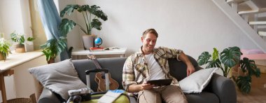 Genç bir adamın resmi, turist tatile çıkıyor, bavulunu hazırlıyor, dijital tabletle kanepeye oturuyor, okuyor, haritasına bakıyor, iş gezisi için kitap oteline gidiyor. Boşluğu kopyala