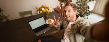 Kafede dizüstü bilgisayarla fotoğraf çeken ve kameraya gülümseyen yakışıklı bir gencin selfie portresi..