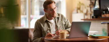 Gözlüklü yakışıklı bir gencin portresi, ders çalışan, not alan, online ders sırasında dizüstü bilgisayara bakan, toplantıya katılan, bilgi yazan, kafede oturan.