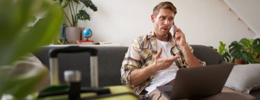 Kızgın bir adamın portresi, hayal kırıklığına uğramış gibi görünüyor, cep telefonuyla konuşuyor, dizüstü bilgisayar ve bavulla oturuyor. Turizm ve halk konsepti