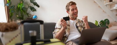 İnternetten bilet alan neşeli bir adamın portresi dizüstü bilgisayar ve kredi kartıyla otel rezervasyonu yapan oturma odasında bavulun yanında oturan....