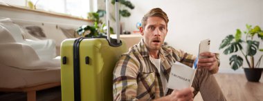 Korkmuş yüzlü bir adam, bavulun yanında oturuyor, akıllı telefon ve pasaport tutuyor. Boşluğu kopyala