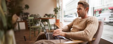 Çalışan insanlar ve kamusal alan kavramı. Genç yakışıklı adam, ux ui tasarımcı, laptopuyla kafede oturuyor, cappuccino içiyor, ciddi bir yüzle ekrana bakıyor..