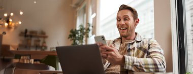 Kafedeki adamın görüntüsü, kafeterya ziyaretçisinin dizüstü bilgisayarı ve akıllı telefonuyla koltukta oturması, cep telefonu ekranına şaşırmış ve heyecanlı görünmesi, büyük haberlere hayran kalması, uygulamanın tanıtım teklifi..