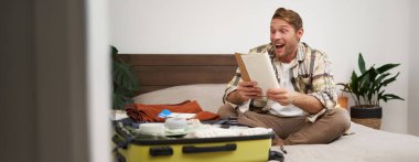 Defterine heyecanla bakan bir adamın portresi, notları okumak, kontrol listelerini gözden geçirmek, tatil için toplanmak, açık bavulun yanında oturmak..