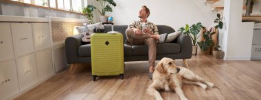 Bavullu, mutlu, yakışıklı genç bir adamın portresi, köpeğiyle oturma odasında oturuyor, tatile çıkmaya hazırlanıyor, tatile çıkıyor. Turizm konsepti