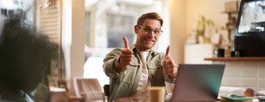 Gözlüklü neşeli güler yüzlü adam, başparmağını kaldırır, laptopla kafede oturur, web sitesi tavsiye eder, e-öğrenim hizmeti veya ortak çalışma alanı.