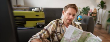 Bavullu mutlu genç adamın portresi, oturma odasında oturur ve haritaya bakar, tatil yerini seçer, seyahat için güzergah seçer..