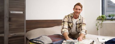 Genç bir adamın portresi, yurtdışına seyahat eden bir turist, bavula giysiler koyuyor, bavulunu kapatıyor, yatağa oturuyor, tatile hazırlanıyor. Turizm konsepti