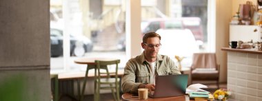 Genç dijital göçebe imajı, gözlüklü adam kafede oturuyor, kahvecide çalışıyor, iş yerinde dizüstü bilgisayar kullanıyor, gözlük takıyor, içkisini içiyor..