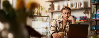 Laptop ekranında hayal kırıklığına uğramış bir adamın portresi, uzaktan çalışan, kafede oturan, kahve içen, keyifsiz, üzgün bir yüz ifadesiyle monitörde mesaj okuyan,.