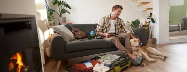 Gülümseyen yakışıklı adam ve köpeği, oturma odasında oturuyor, yaz tatili için bavul hazırlıyor, bir tatil planlıyor, elinde defter tutuyor..