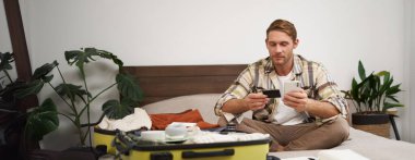 Genç bir adamın portresi, internetten ödeme yapan bir turist, kredi kartı ve cep telefonu uygulaması kullanıyor, bavulla yatakta oturuyor, internetten sipariş veriyor, tatil biletleri alıyor..