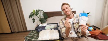 Kulaklıklı yakışıklı bir adamın portresi, açık bavuluyla yatakta oturuyor, iki uçak biletiyle pasaportunu gösteriyor, seyahat acentesini tavsiye ediyor, tatile çıkıyor, tatil için heyecanlanıyor..