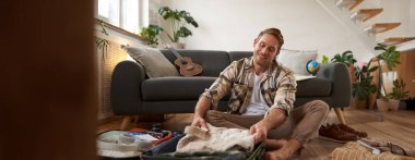 Seyahat ve turizm konsepti. Mutlu genç adam, bagajı açık bir şekilde yerde oturuyor, tatil için bavulunu hazırlıyor, tatile çıkıyor, kıyafetlerini bir çantaya koyuyor..