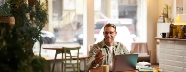 Gözlüklü yakışıklı bir adamın portresi, laptopla kafede oturuyor, telefonda mesaj okurken gülümsüyor, akıllı telefon uygulamasını kontrol ediyor, kahve dükkanından çalışıyor..
