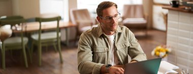 Gözlüklü genç bir adamın portresi, kafede çalışıyor, ders çalışıyor, laptoptaki görevi tamamlarken düşünceli görünüyor, not defterine notlar alıyor..