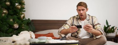 Şok olmuş bir adamın portresi, akıllı telefonuna ve kredi kartına bakıyor, bavuluyla oturuyor, tatil için hazırlanıyor, hesap bakiyesi için endişeli görünüyor..