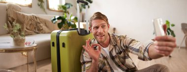 Sosyal medyada tatille ilgili yakışıklı turist gönderileri, tamam işareti ve bavuluyla selfie çekiyor, akıllı telefon uygulaması kullanıyor. Turizm ve tatil kavramı